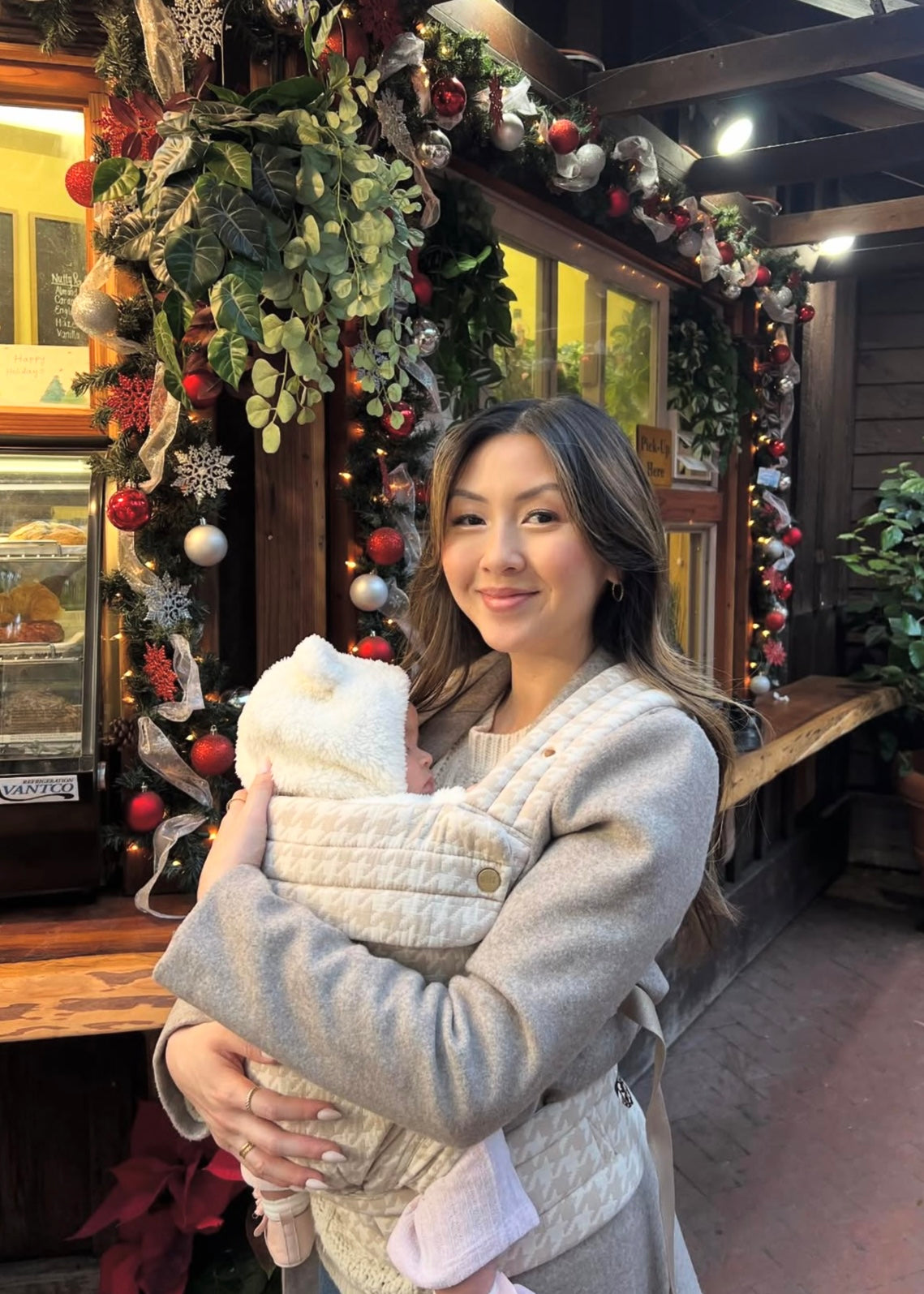 Woman carrying baby in quilted beige baby carrier, festive holiday decorations in background