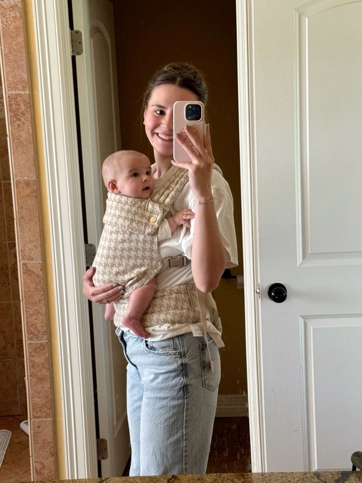 Mother holding baby in beige patterned baby carrier in front of bathroom mirror