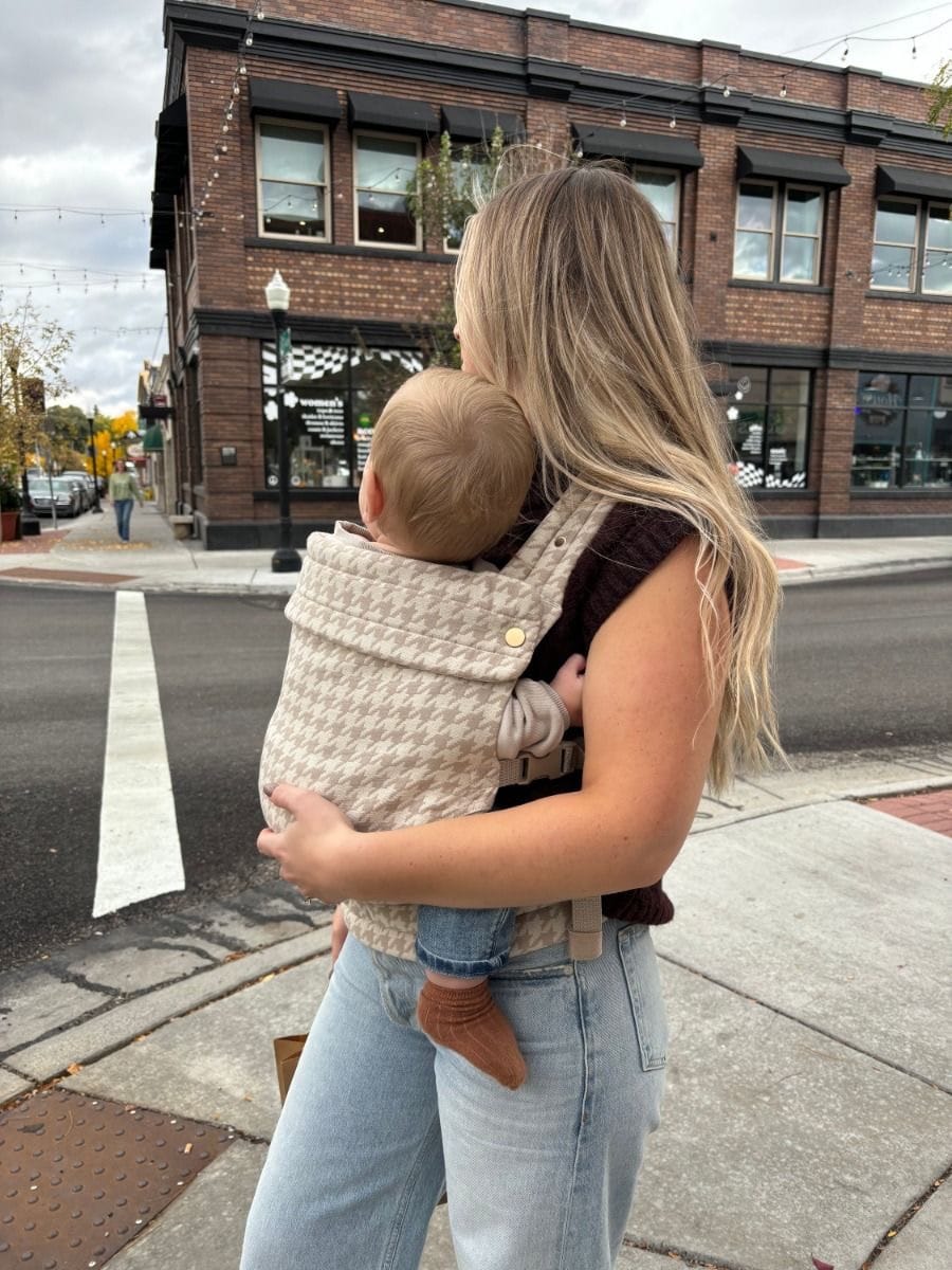 Woman carrying baby in beige houndstooth baby carrier on city street corner