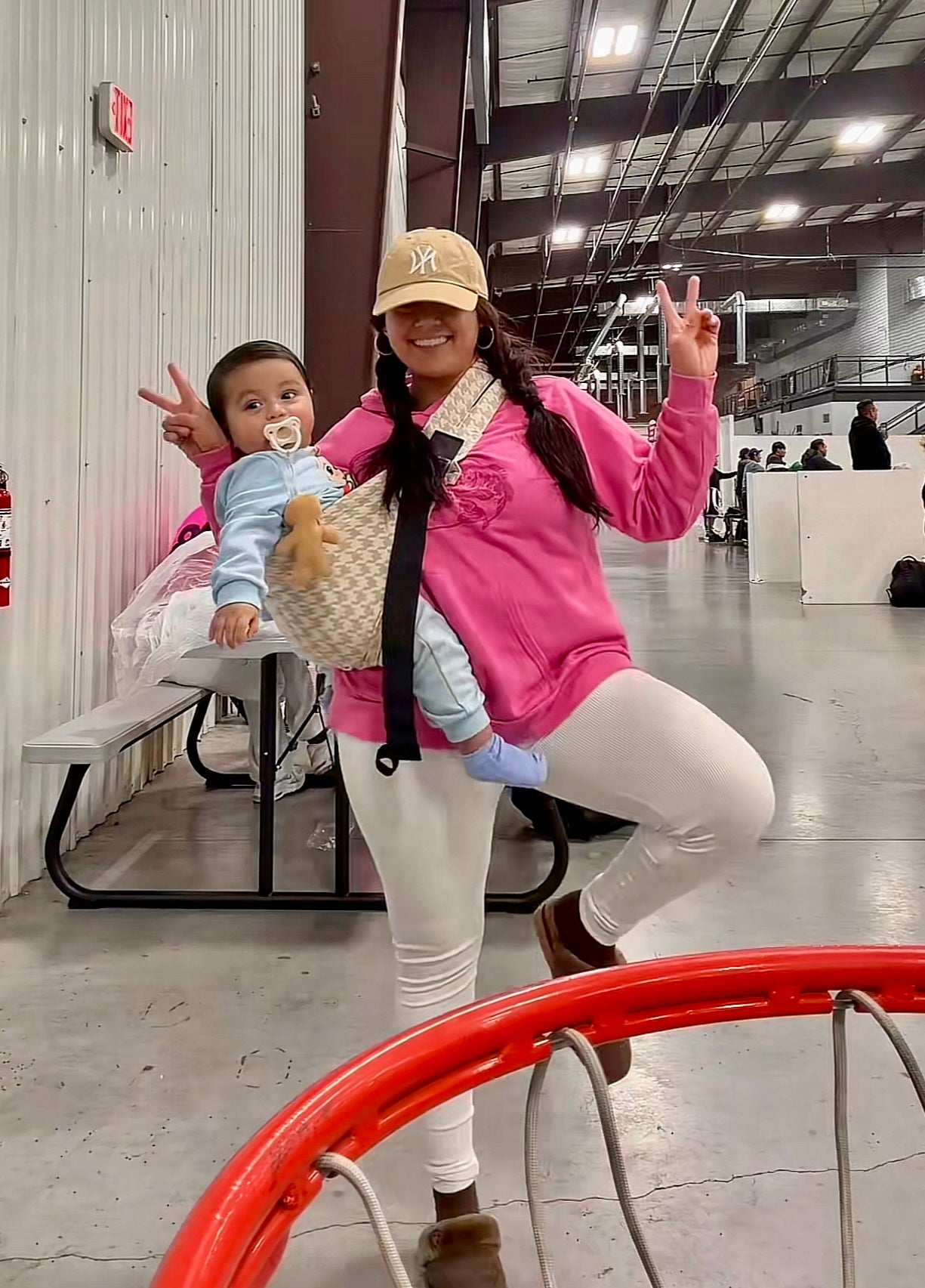 Woman wearing pink hoodie poses with baby in beige baby carrier at indoor sports facility