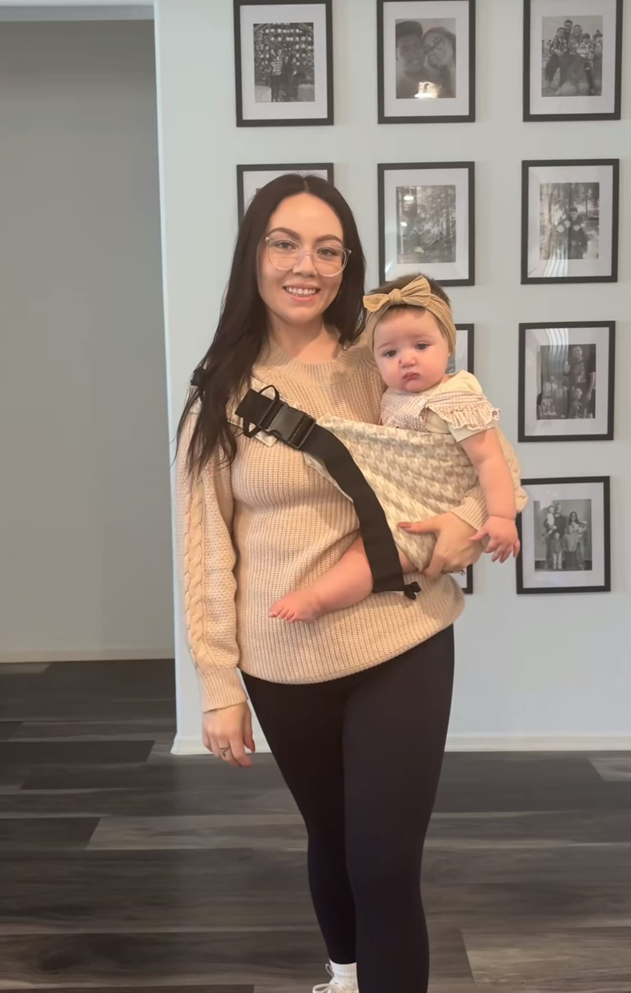 Woman wearing glasses holds baby in patterned baby carrier sling indoors