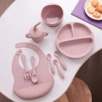 Pink silicone baby feeding set with bib, utensils, divided plate, bowl, and sippy cup on table