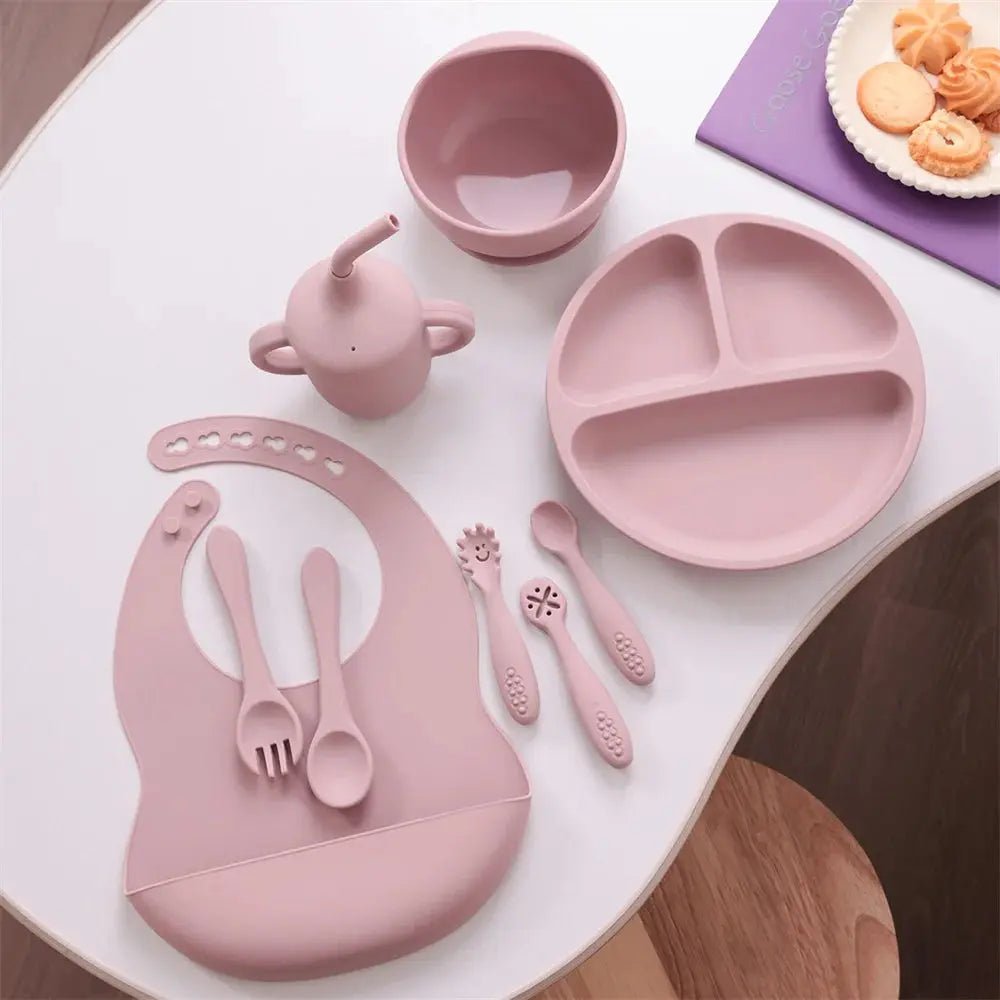 Pink silicone baby feeding set with bib, utensils, divided plate, bowl, and sippy cup on table