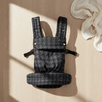 Black houndstooth baby carrier with padded straps on light wood floor, soft white cloth nearby