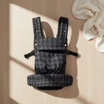 Black houndstooth baby carrier with padded straps on light wood floor, soft white cloth nearby