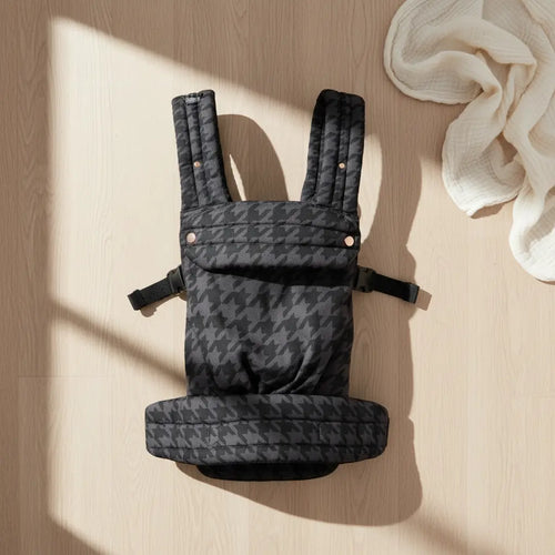 Black houndstooth baby carrier with padded straps on light wood floor, soft white cloth nearby